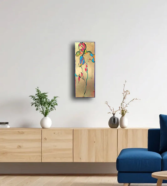 Ein goldfarbenes Gemälde im Hochformat mit einer Lilie die rote und blaue Blüten trägt. An einer hellen Wand hängend über einem Sideboard.