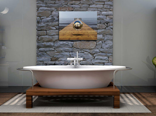 Eine Kugel schwebt über einem Steg der in die Ostsee führt-Surreales Werk, an einer Steinwand über eine Badewanne hängend.